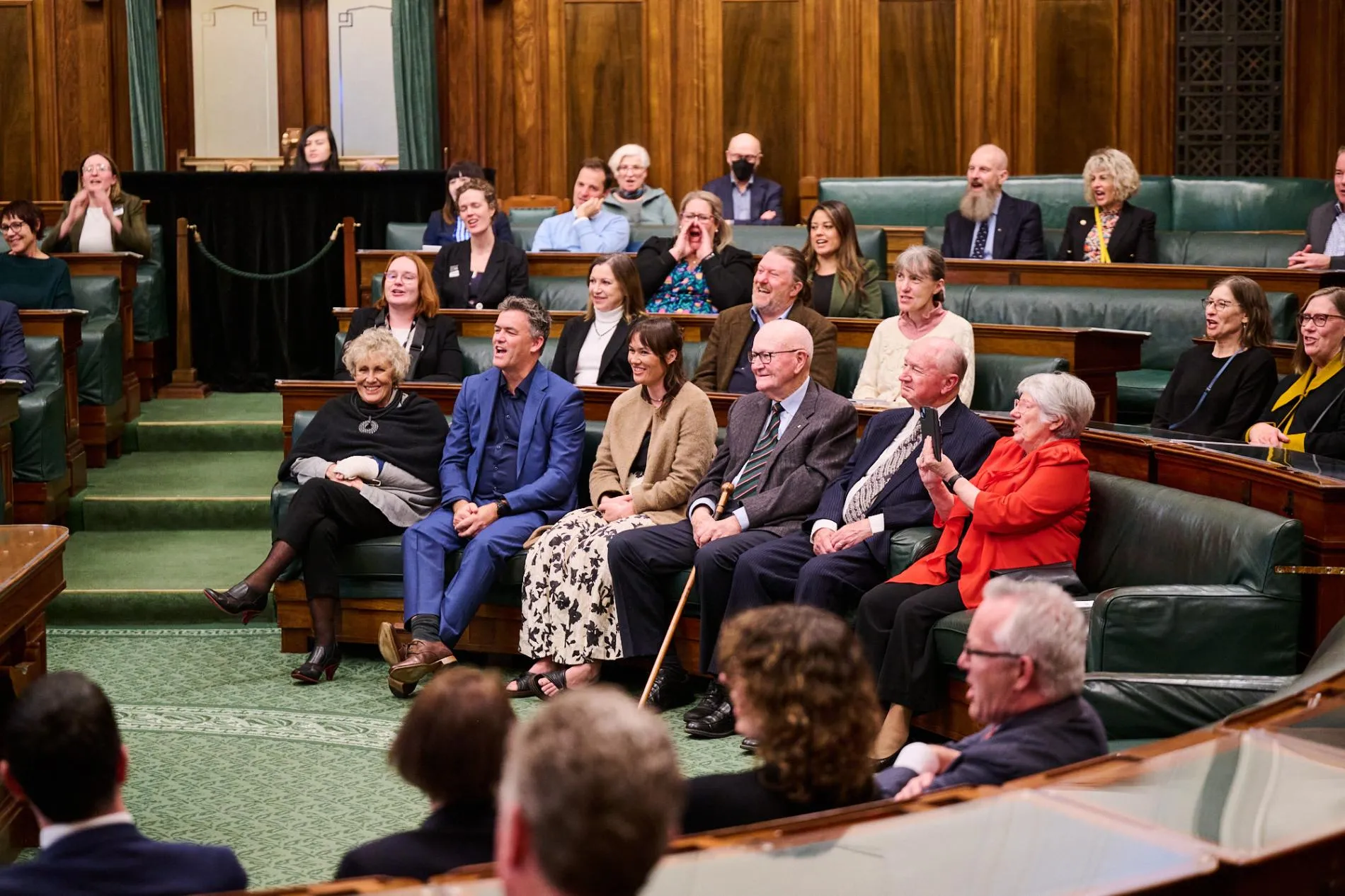 A group of people sit in the House of Representatives Chamber at Old Parliament House, it has green carpet.