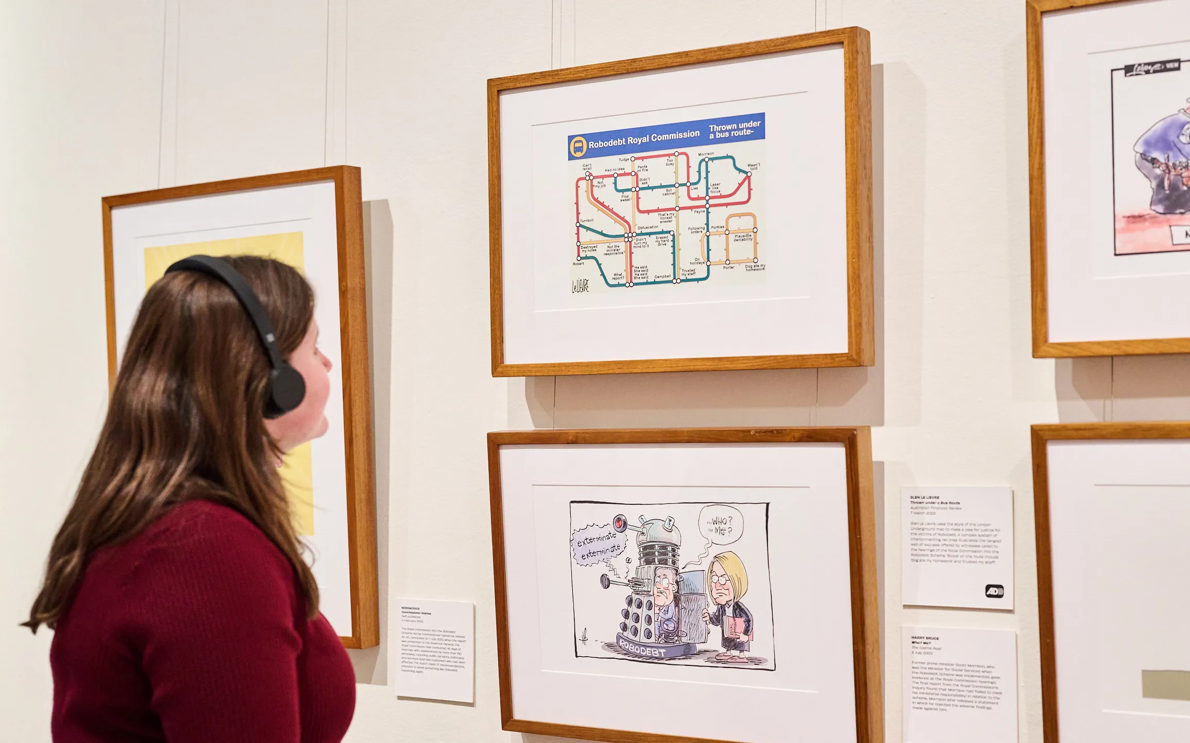 A woman wearing a dark red top and black headphones looks at framed cartoons with a caption featuring the Audio Description logo.