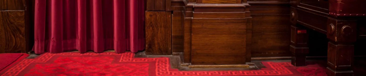 Close up of a red velvet curtin falling to a red carpeted floor with a timber furniture panel next to it.