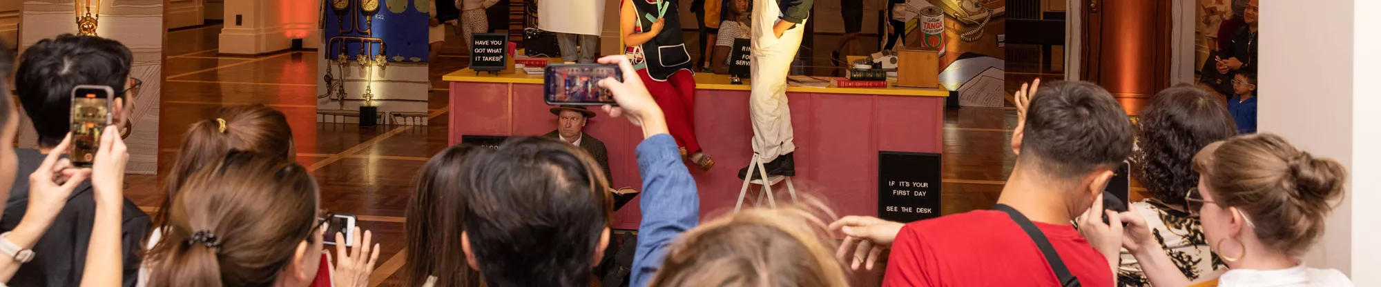 A group of visitors hold up their phones and film a performance with actors dressed as a chef, a hairdresser and a handyman inside King's Hall with a sign in the background that says The People's House. 