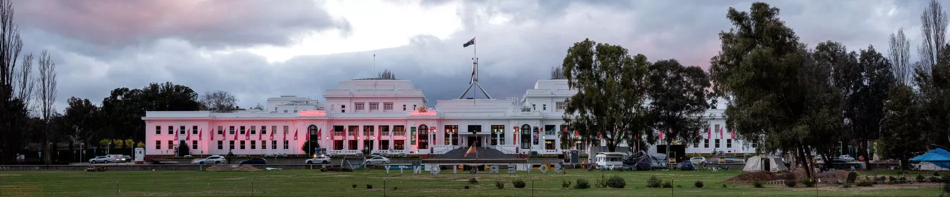 A large white building with the Australian flag on top, with a body of water in front that shows the reflection. The image is at dusk. 