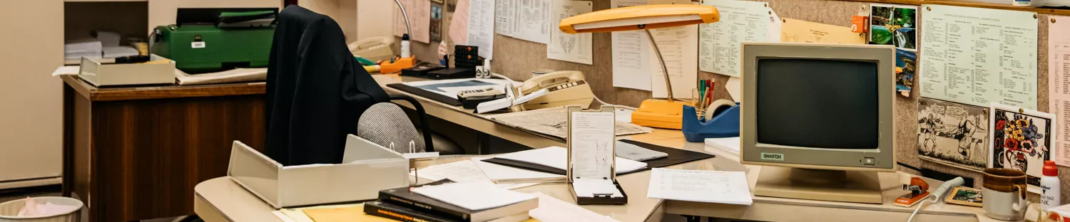 Two desks cluttered with books, pens, staplers, old fashioned 80s computers, and a pinboard covered in papers. 