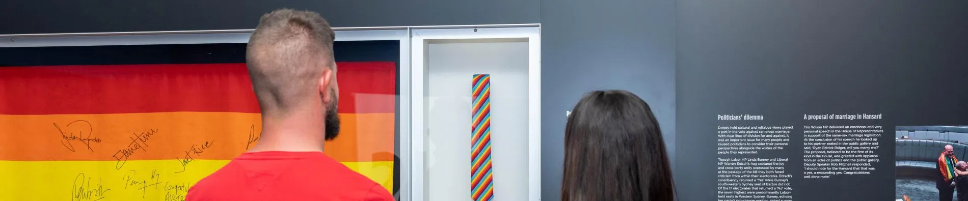 A man and a woman stand in front of a display case with a rainbow flag with signatures on it. 