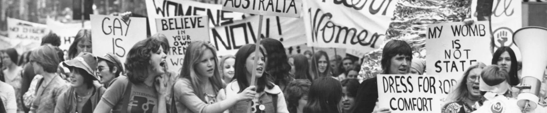 A black and white photo of a group of women holding protest signs in 1975. Signs say 'sexism is alive and well in Australia' and 'dress for comfort not style'.