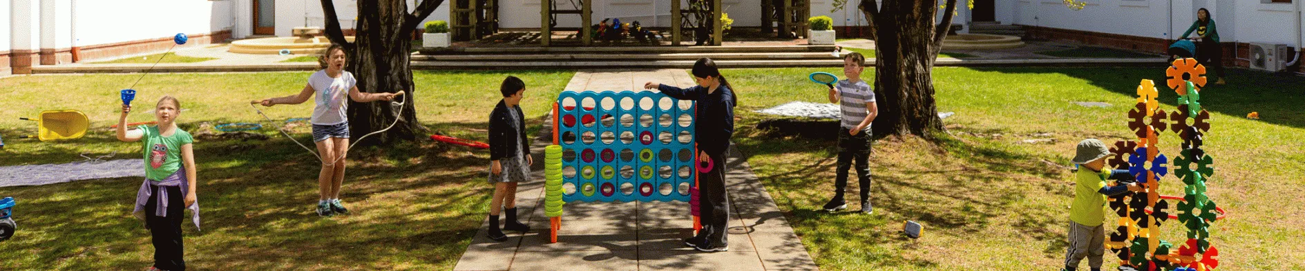 Children play in a green courtyard surrounded by trees and the white walls of Old Parliament House. There is a large connect 4 game, hula hoops and a skipping rope.