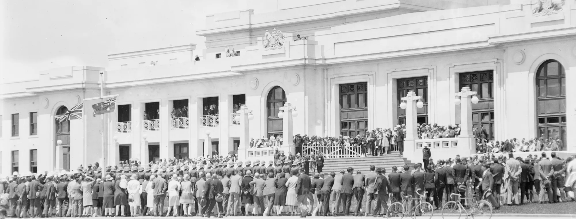 Black and white image of the front of Old Parliament House with a crowd of people of gathered at the foot of the stairs. The building is white with columns and an Australian flag flies at the top. 