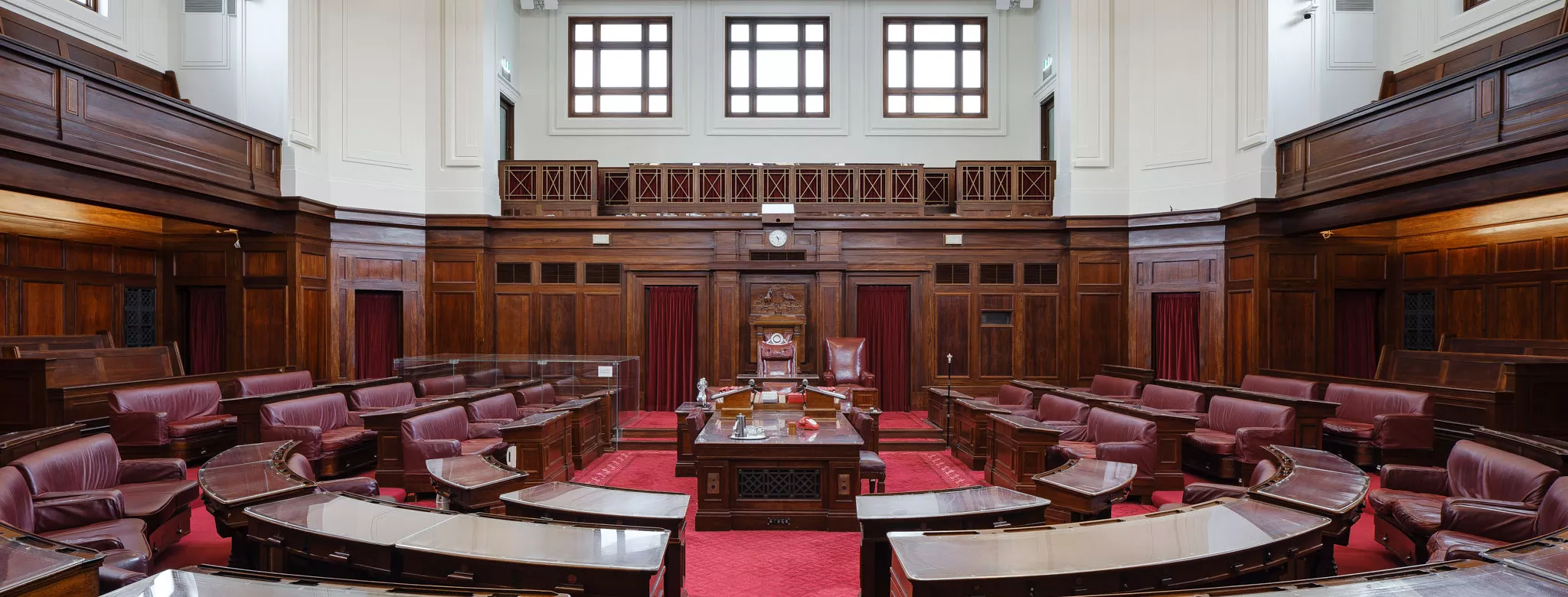 A view into the Senate Chamber looking straight into the room with benches in a U-shape on either side of the centre table. The carpet is a rich red and the seats are red leather. Art deco lights hang from the ceiling and wood timber panels surround the room. 