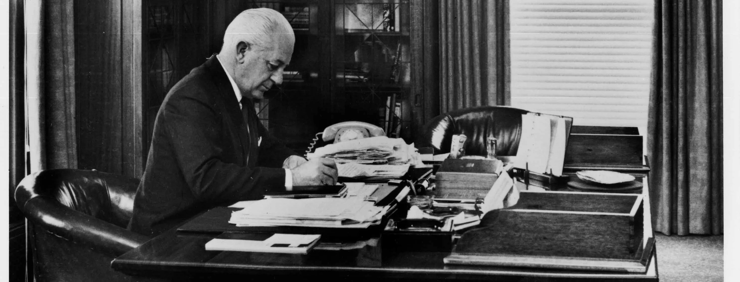 In this black and white photograph Prime Minister Harrold Holt sits at the timber desk made in 1926. Holt is wearing a dark suit, white shirt and tie and is leaning on the desk while he writes. The desk has fine carved panels with a geometric detailing on the corners. The surface of the desk is cluttered with stacks of documents and a telephone. On the glass-fronted cabinets in the background are a number of framed photographic portraits.  