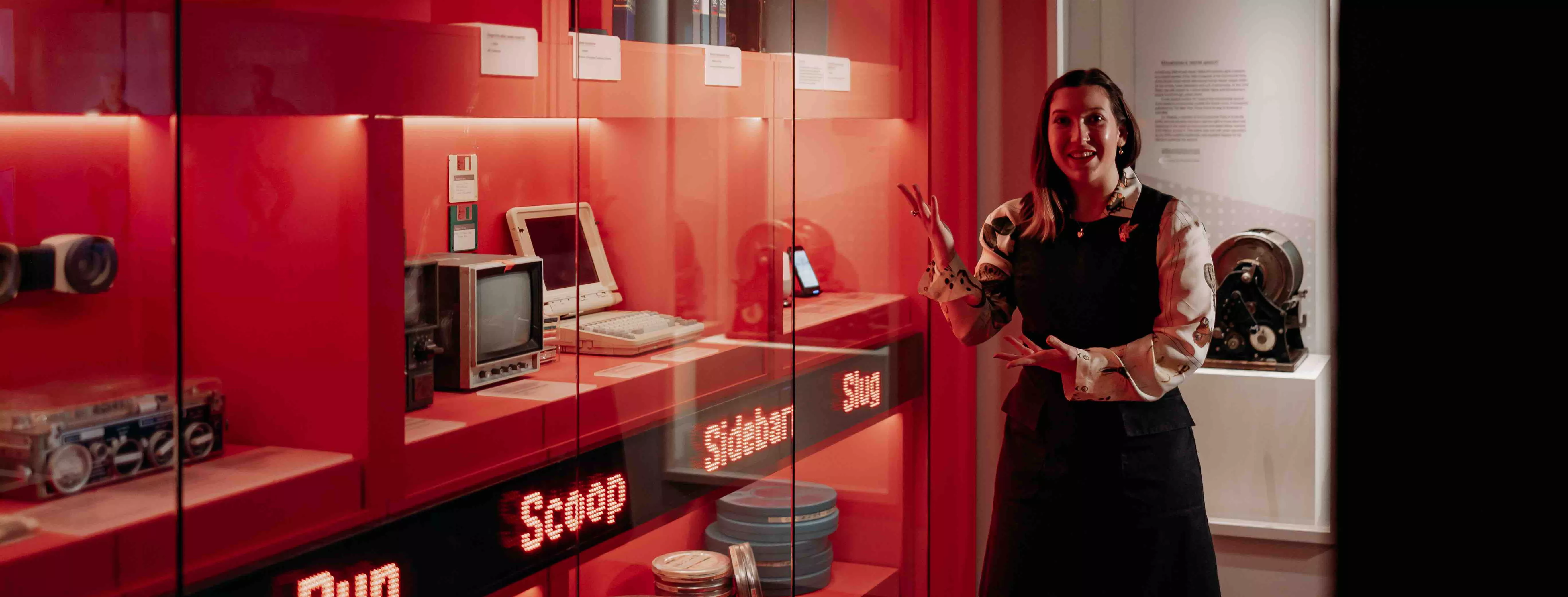 A MoAD educator stands in front of the Truth, Power and a Free Press exhibition wall featuring red walls with different types of media technology behind the glass case. 