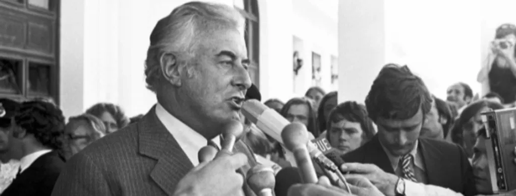 Black and white photograph of Gough Whitlam speaking to a large group of people holding microphones to his face, in front of parliament house immediately following the Dismissal.