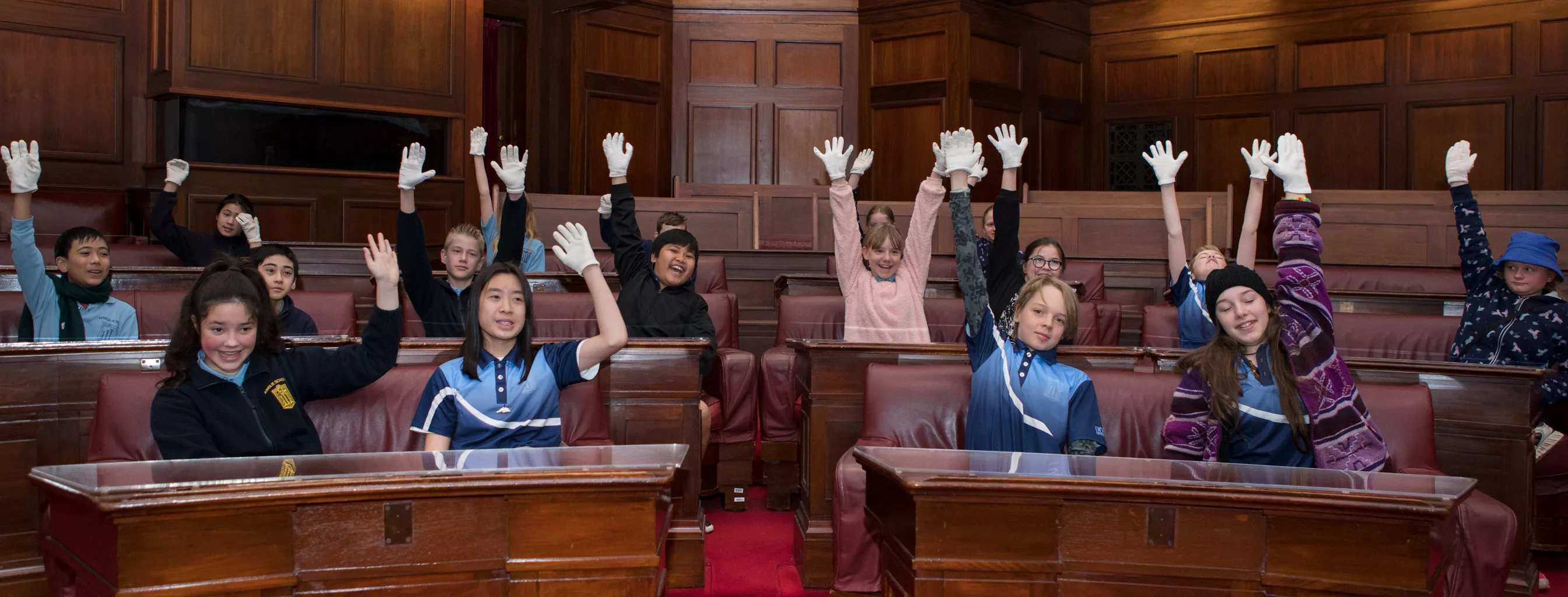 School children raise their hands while sitting in the Senate Chamber which has red carpet and wood panelled benches in a U-shape. 