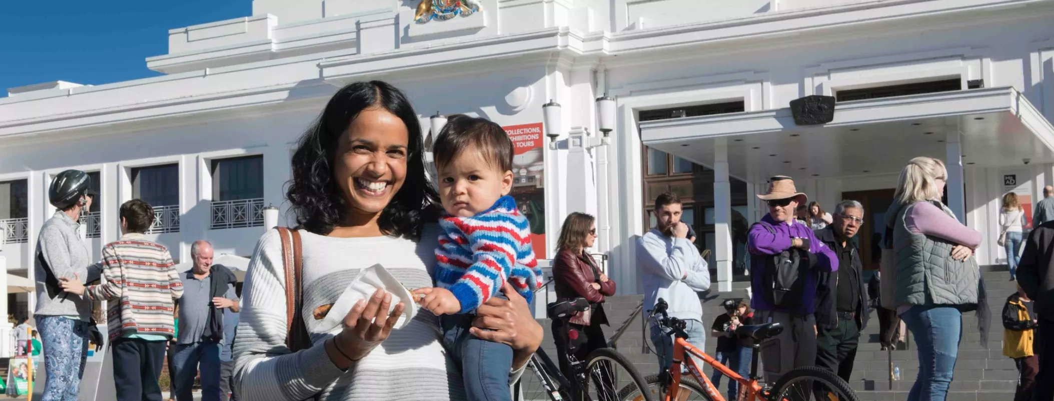 A mother holds her baby and smiles at the camera at the front of Old Parliament House. A row of people waiting to vote are behind her. She holds a sausage sandwich.