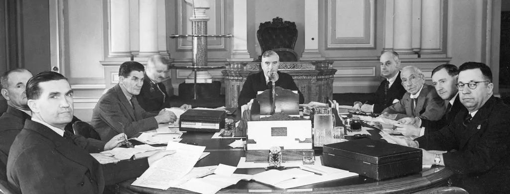 Nine men are gathered, seated around a large table covered with paper documents, with Robert Menzies seated at the head of the table.