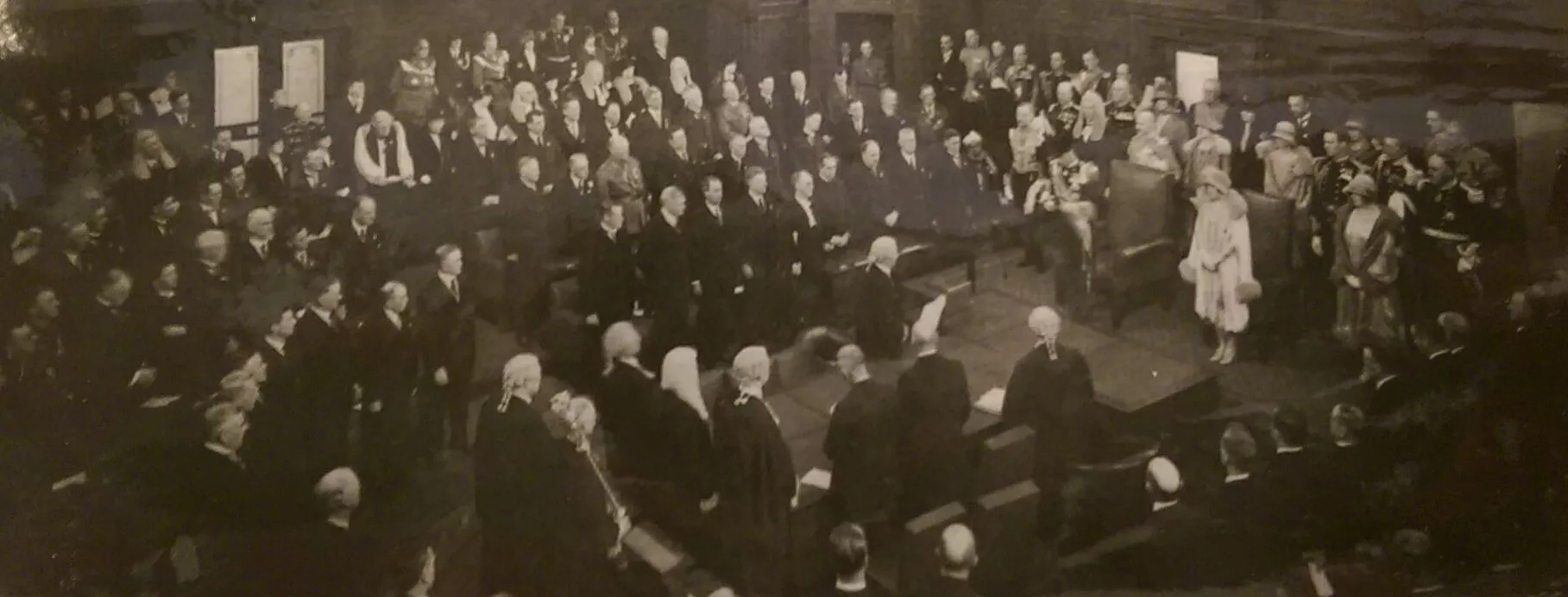 This black and white photograph shows the Duke of York reading a message from his father the King in the Senate Chamber. The room is crowded with at least 200 people, mostly men with some women all wearing their best. Men are dressed in suits, military uniform or gowns and wigs while the women wear formal dresses with hats and gloves. The crowd is in a horseshoe shape around the central table while the Duke stands at the head of the table.