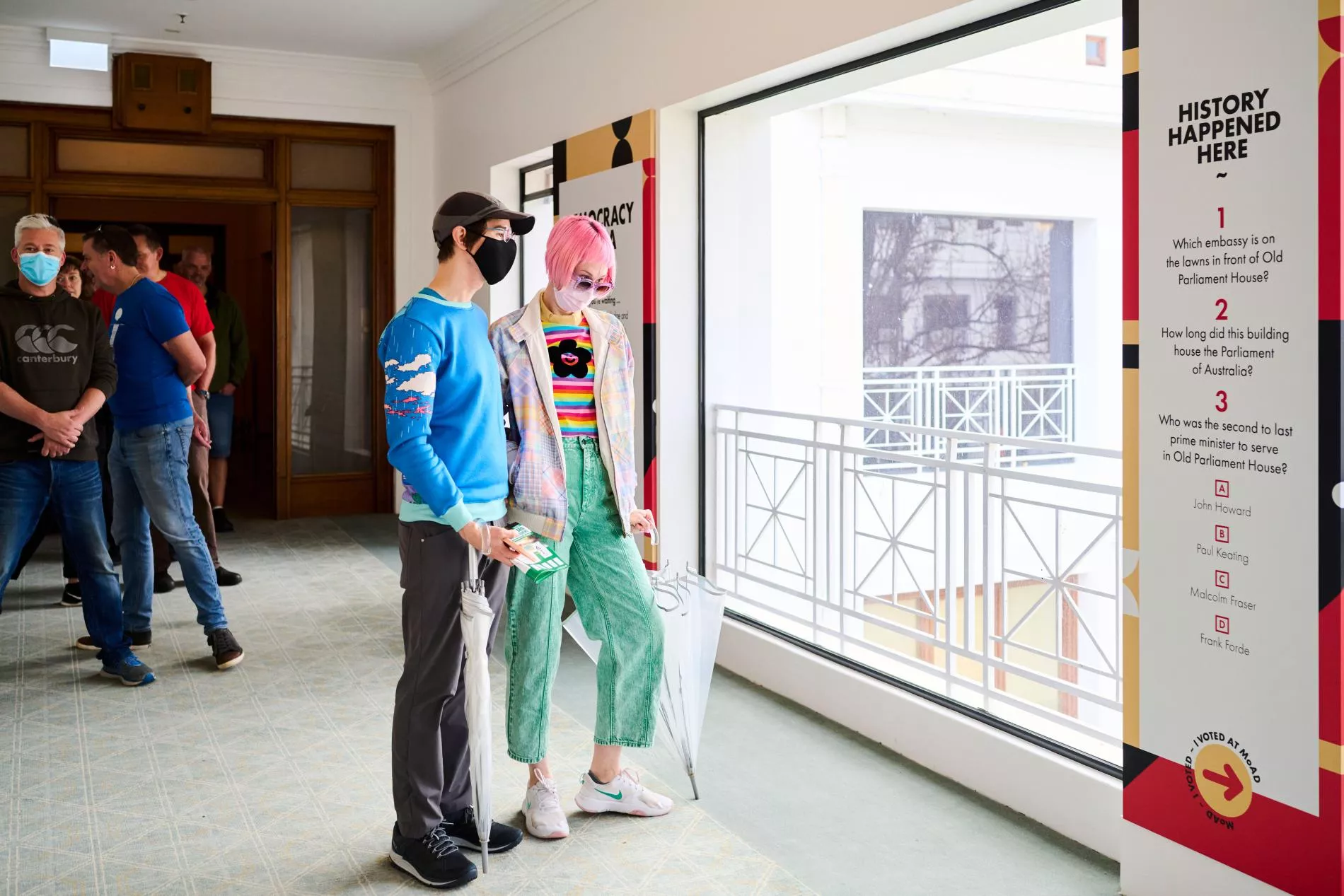 A man and a woman look at a sign inside the museum. The girl has pink hair and a rainbow shirt and wears a face mask. The man wears a blue shirt and a face mask.