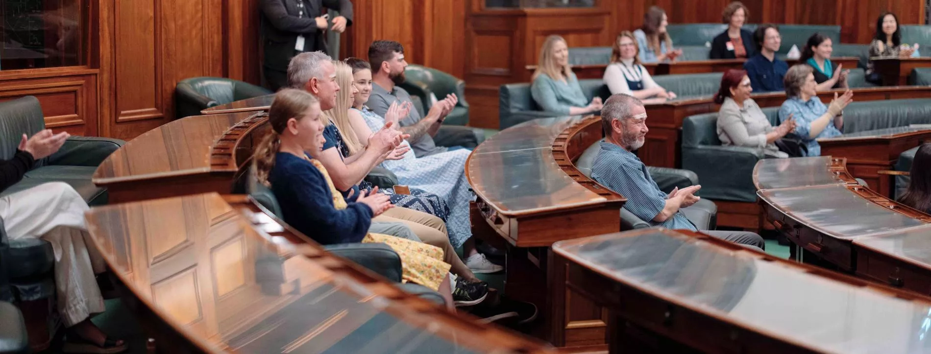 Visitors sit in green leather chairs with timber desks with glass coverings in a U-shape in the House of Representatives. 