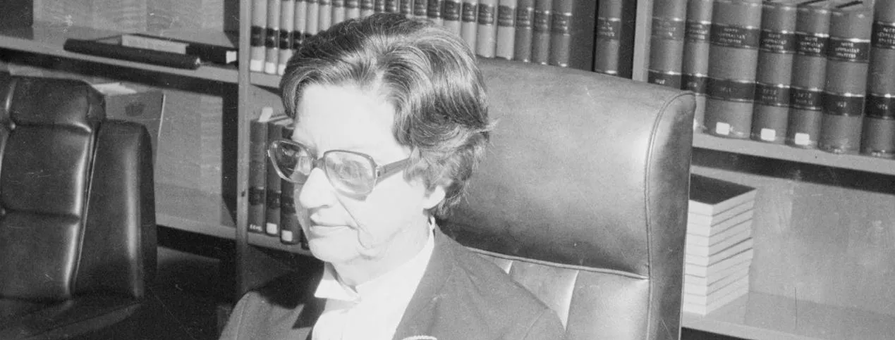 A woman, Justice Roma Mitchell sits at a desk covered in papers, in front of a bookshelf filled with bound volumes.