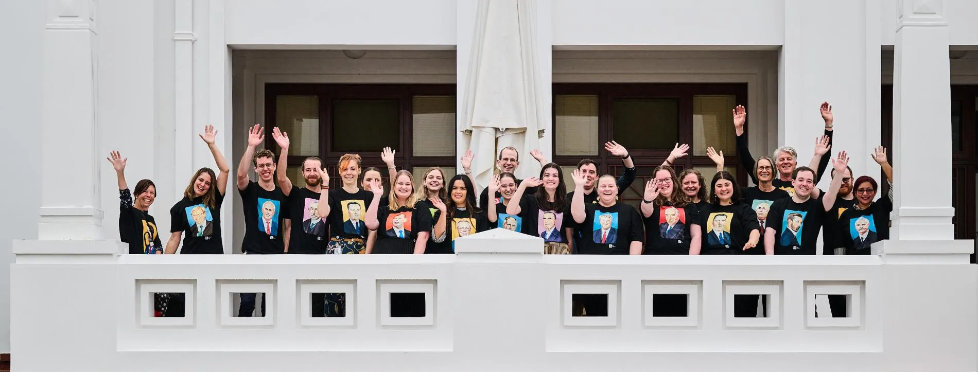 A group of people in black T-shirts with illustrations of prime ministers on them wave from a white terrace balcony.