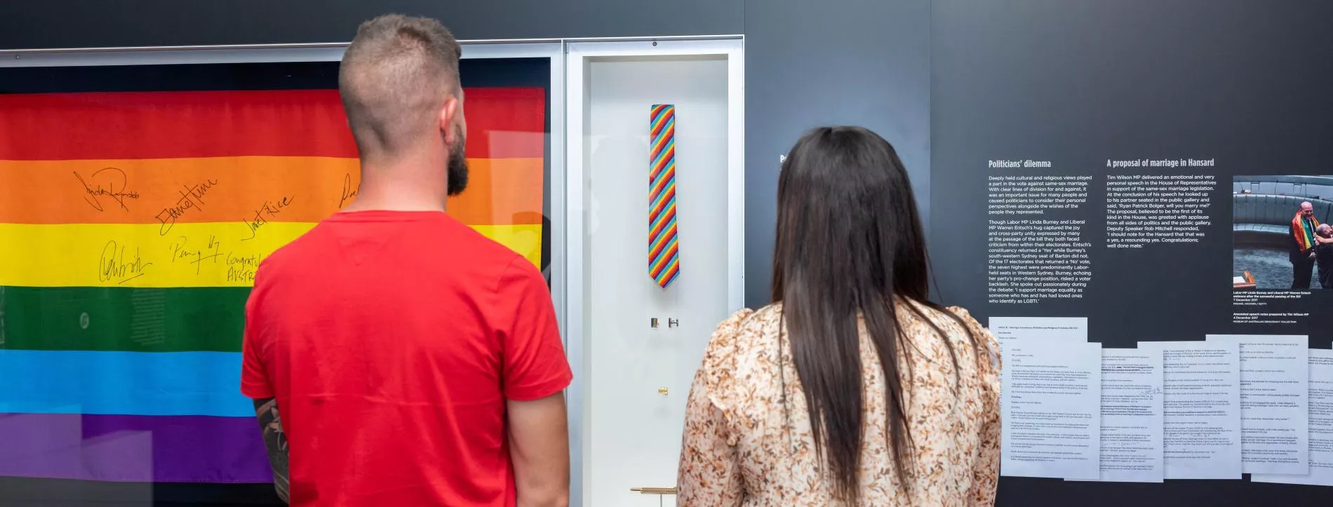 A man and a woman stand in front of a display case with a rainbow flag with signatures on it. 