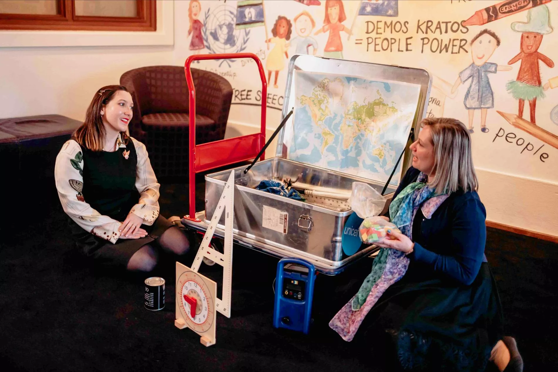 Two MoAD educators sit on the floor with a large metal storage box between them with a map and other objects inside.