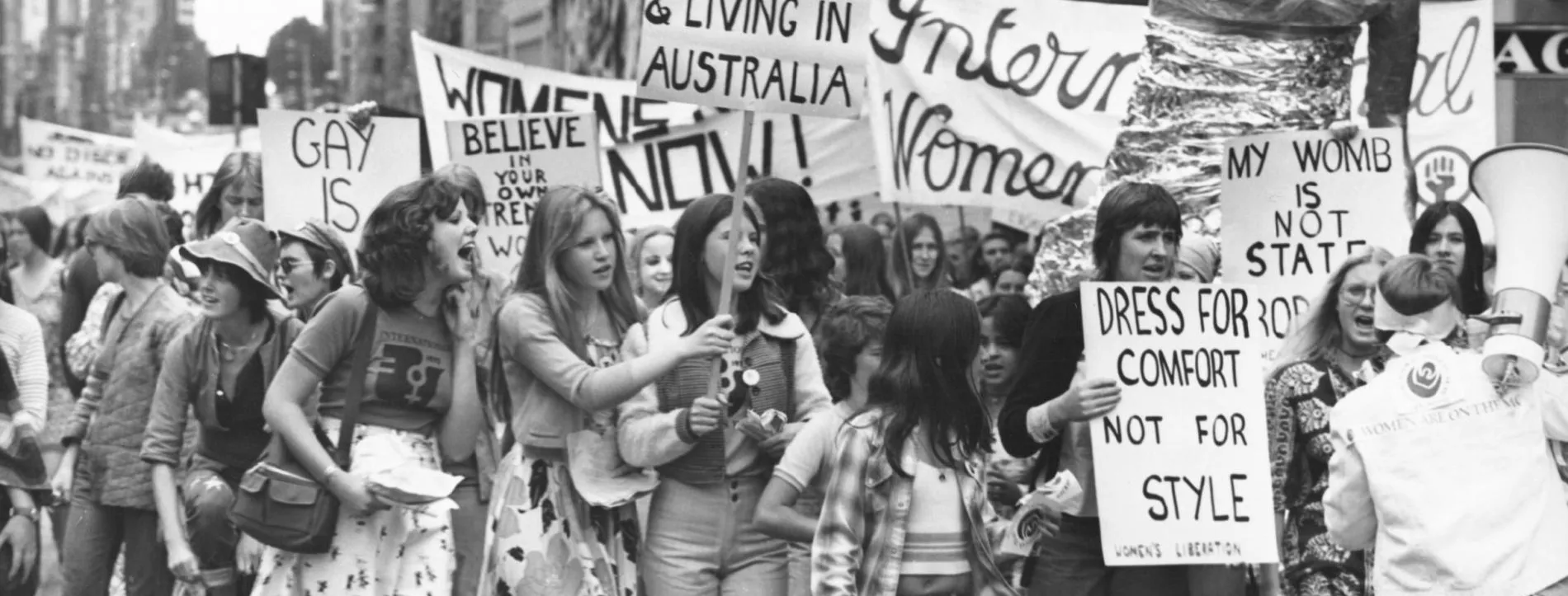 A black and white photo of a group of women holding protest signs in 1975. Signs say 'sexism is alive and well in Australia' and 'dress for comfort not style'.