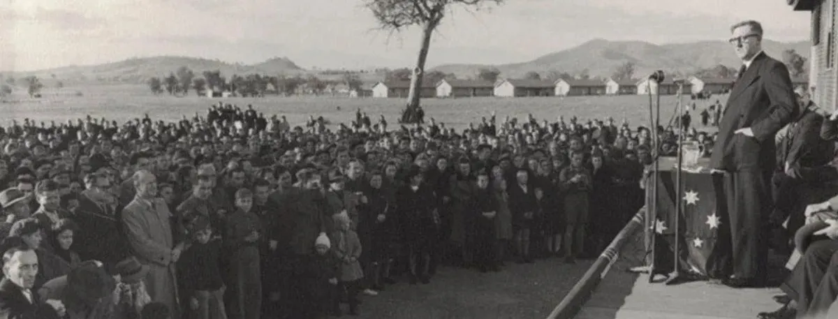 Arthur Calwell stands on a platform with microphones, addressing a crowd of people. In the distance is a row of long buildings.