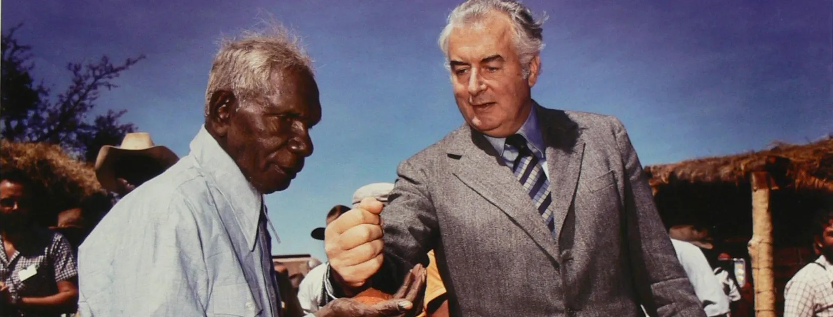 Prime Minister Gough Whitlam pouring earth through Vincent Lingiari’s hand while people look on.