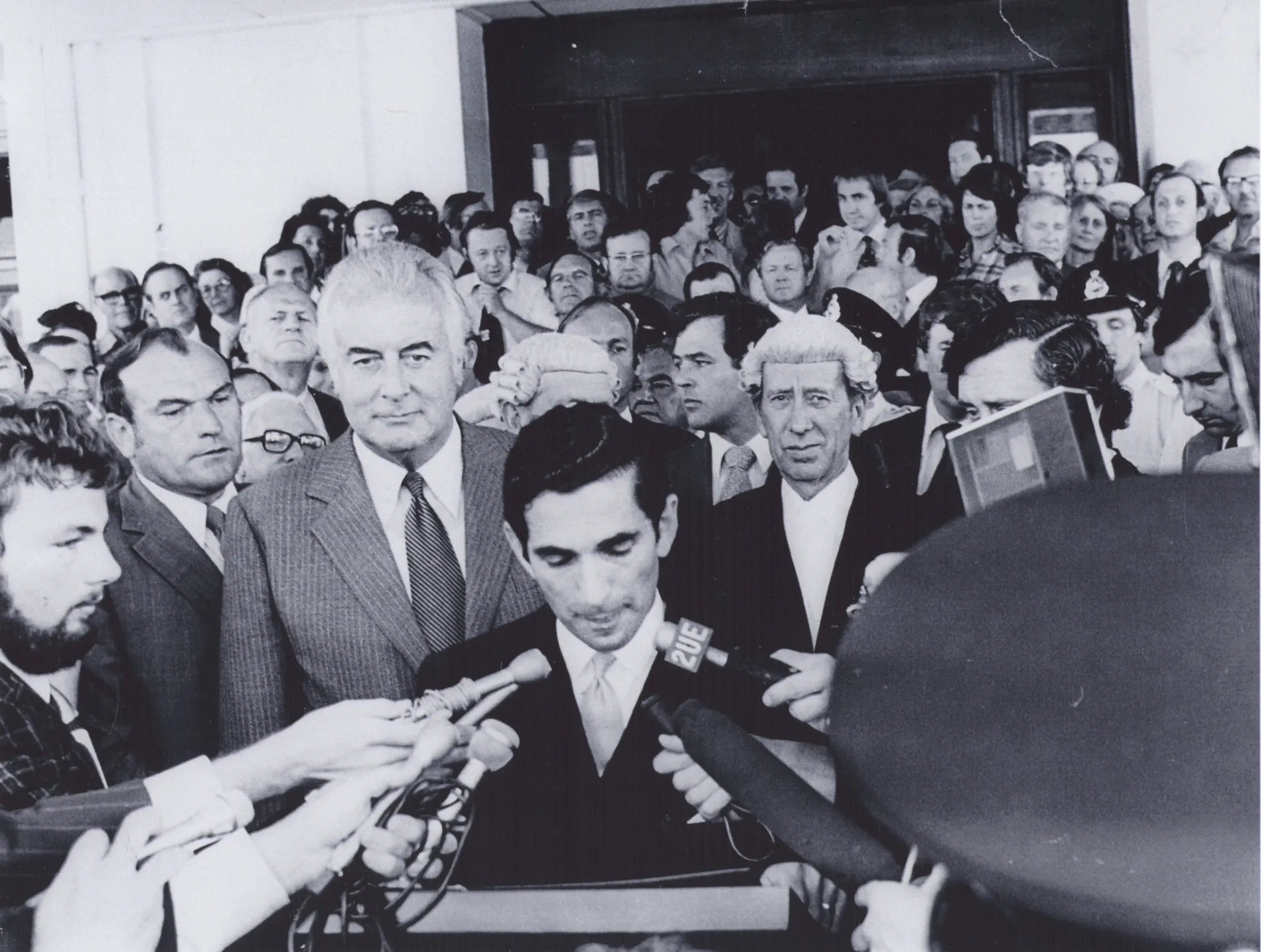A black and white photo of a Gough Whitlam on the steps of Parliament House surrounded by a crowd of men in suits. One many at the front speak from a lecturn as journalists put their microphones in his face. A man in the background wears barrister's wig and looks at the camera. 