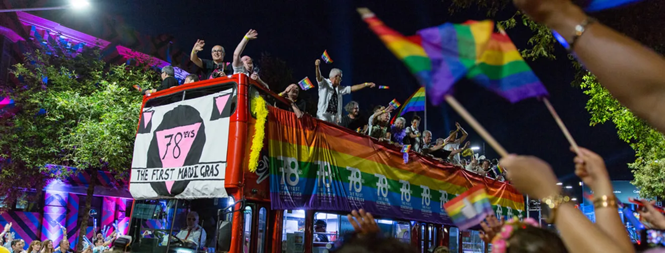 A red double-decker bus with rainbow banners displaying the words '78ers: the first mardi gras' with people on top of the bus waving to crowds on each side of the street.