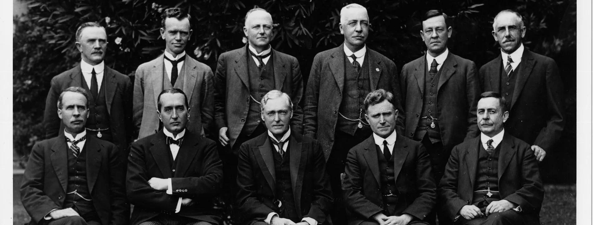 Black and white photograph of eleven men, posing in two rows, all wearing three-piece suits.