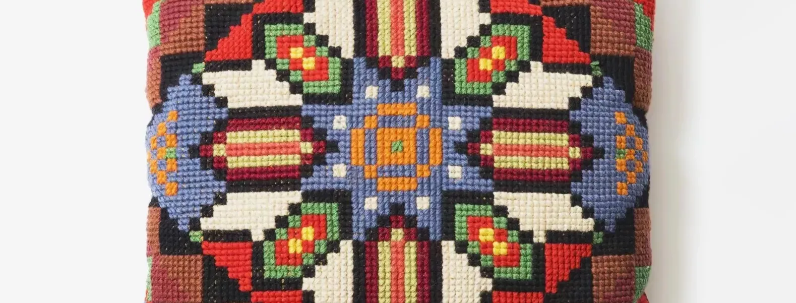 A square embroidered cushion with a geometric pattern using green, yellow, red, blue, orange, black and white threads.