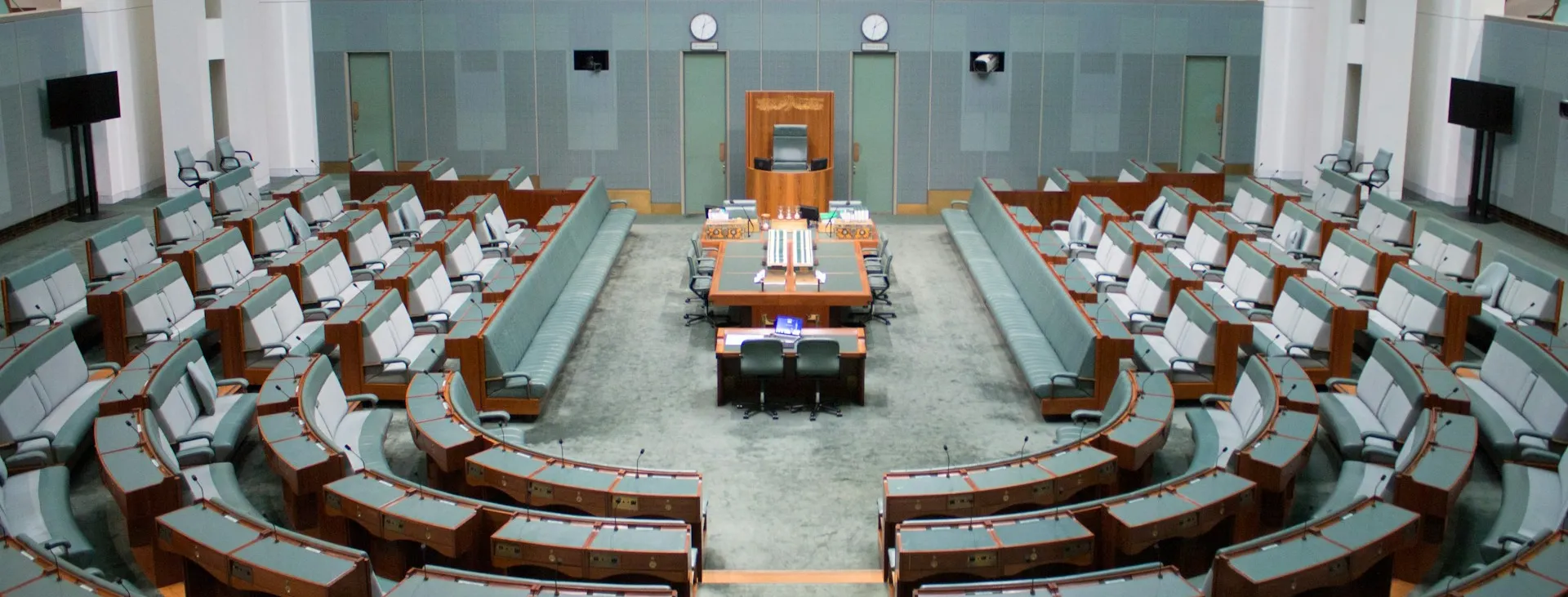 The House of Representatives, a room with grey-green furnishings, with benches arranged in a horse-shoe shape around a central long bench and chair.