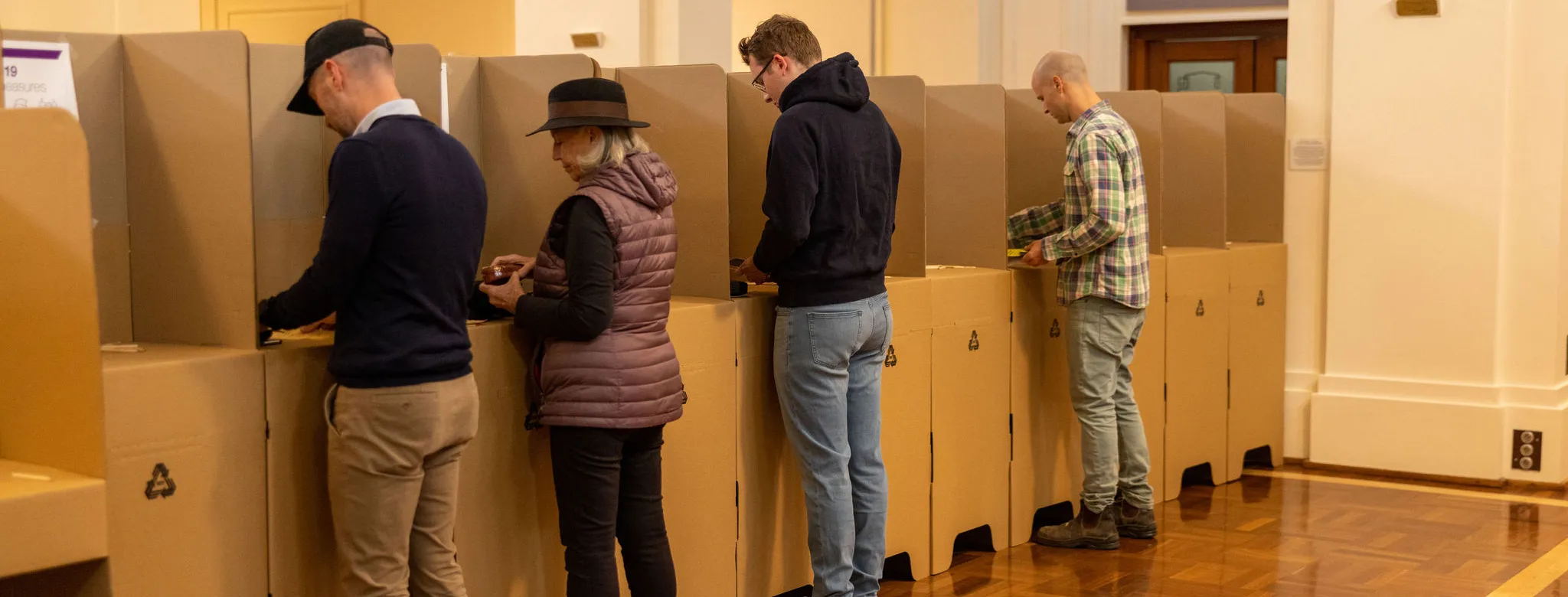 Four people, three men and a woman, standing at cardboard election booths in King's Hall.