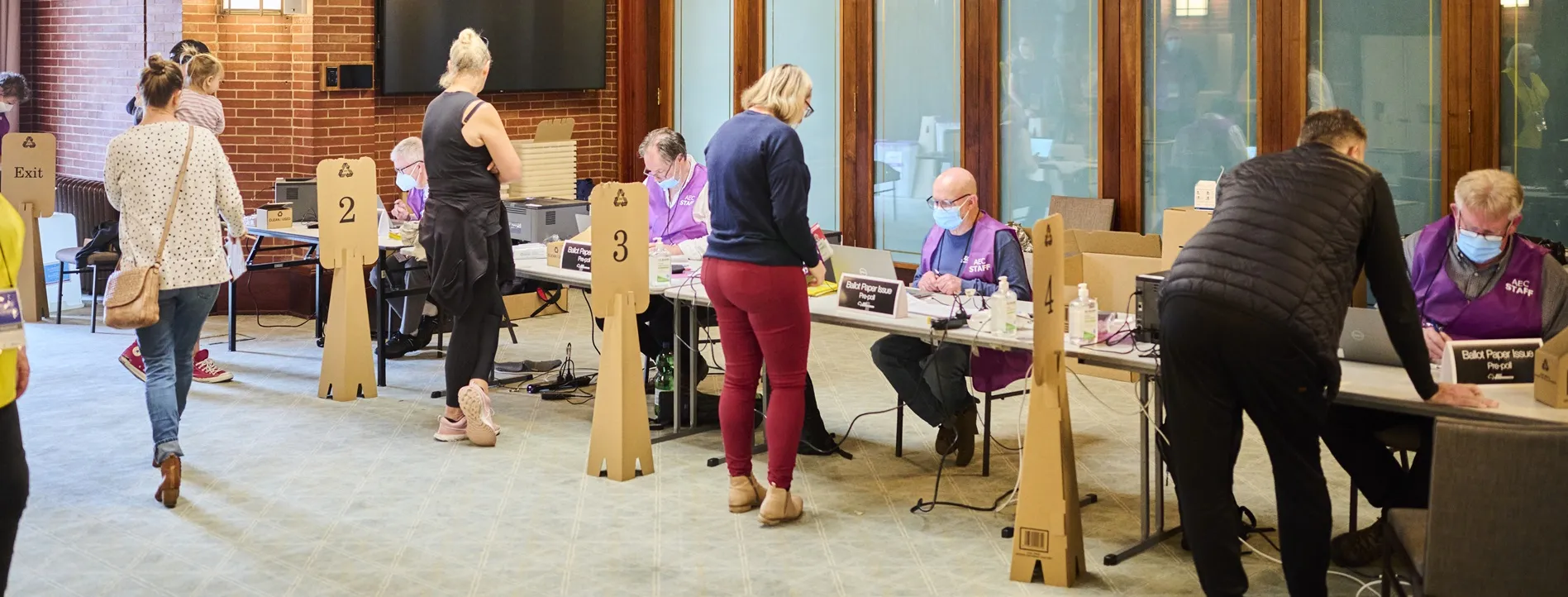 Four Australian Electorial Commission staff sit in a row at a long table wearing face masks and purple vests, and people are lined up, one in front of each staff member, to collect their ballots.