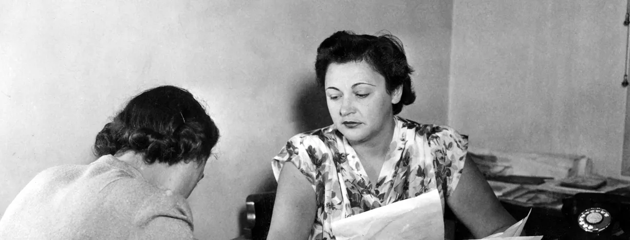 Nancy Wake is at a desk holding papers. She is looking across at another woman who is writing.