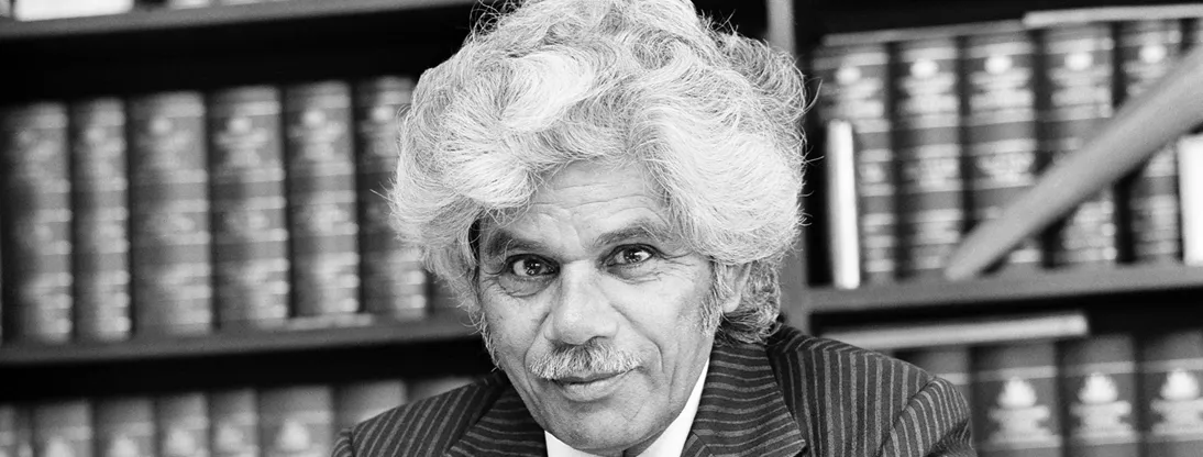 Neville Bonner is sitting at his desk wearing a suit, holding a pen. There are books on bookshelves behind him.