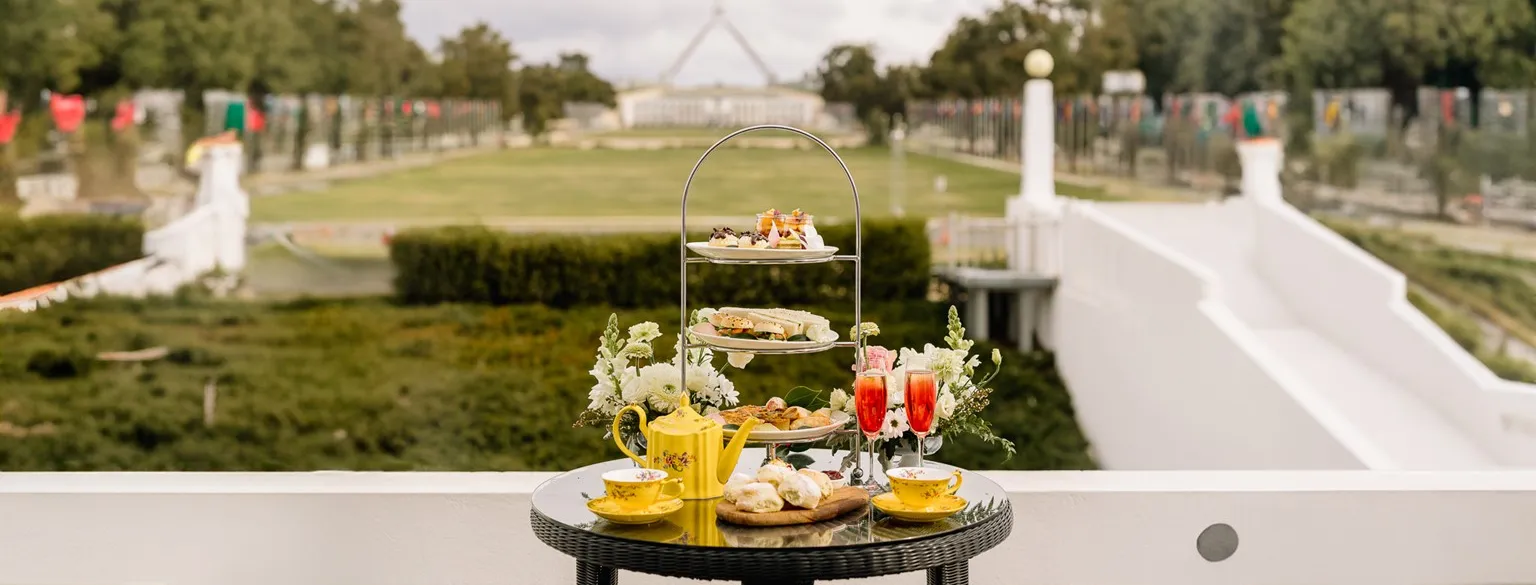 A table is set with a high tea tower filled with cakes and sandwiches is on the balcony of Old Parliament House. Australian Parliament House is in the background along with green lawns.