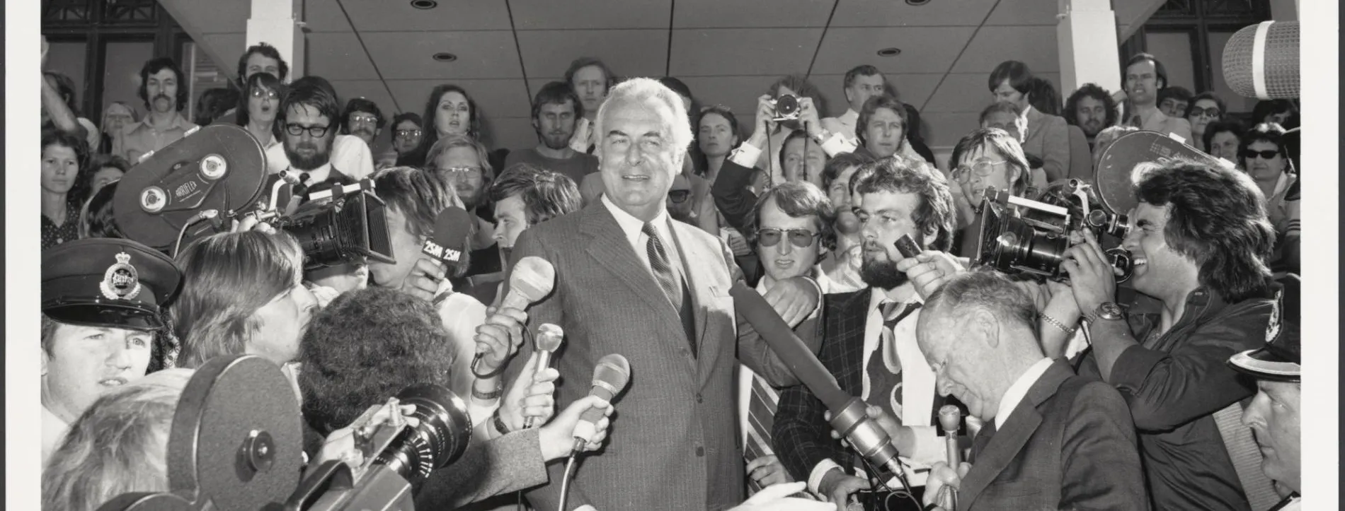 Gough Whitlam on the steps of Old Parliament House surrounded by the press who are holding microphones and video cameras on 11 November 1975.