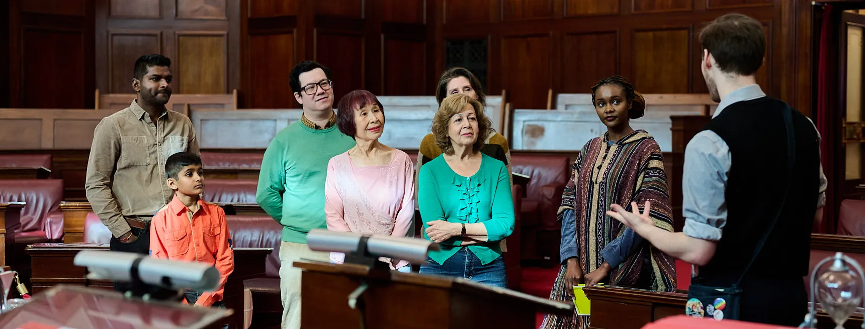 A tour guide faces a group of people who listen to what he is saying. They are standing in front of the red leather bench chairs of the Senate Chamber.