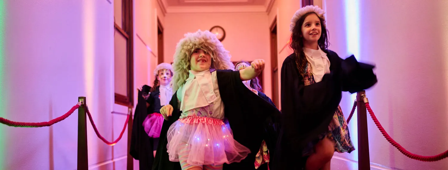 Two girls dressed up in costume white wigs and black gowns walk down a hallway with red carpet in Old Parliament House. One of the girls is also wearing a pink tutu with LED lights in it.