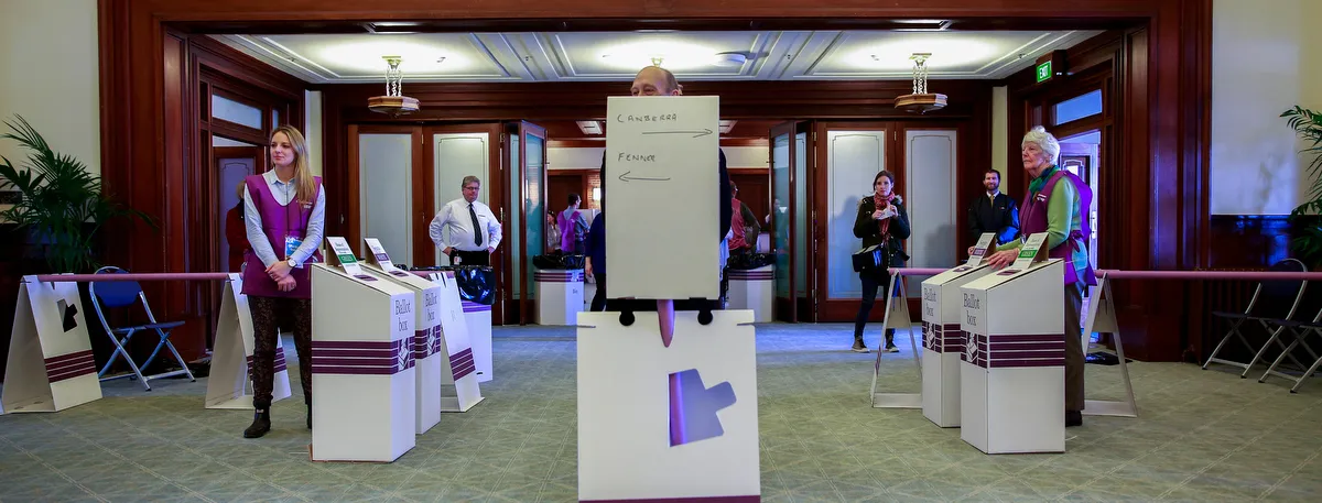 White ballot boxes for Canberra and Fenner are set up on green carpet in Old Parliament House. Australian Electoral Commission staff in purple vests stand next to the boxes.