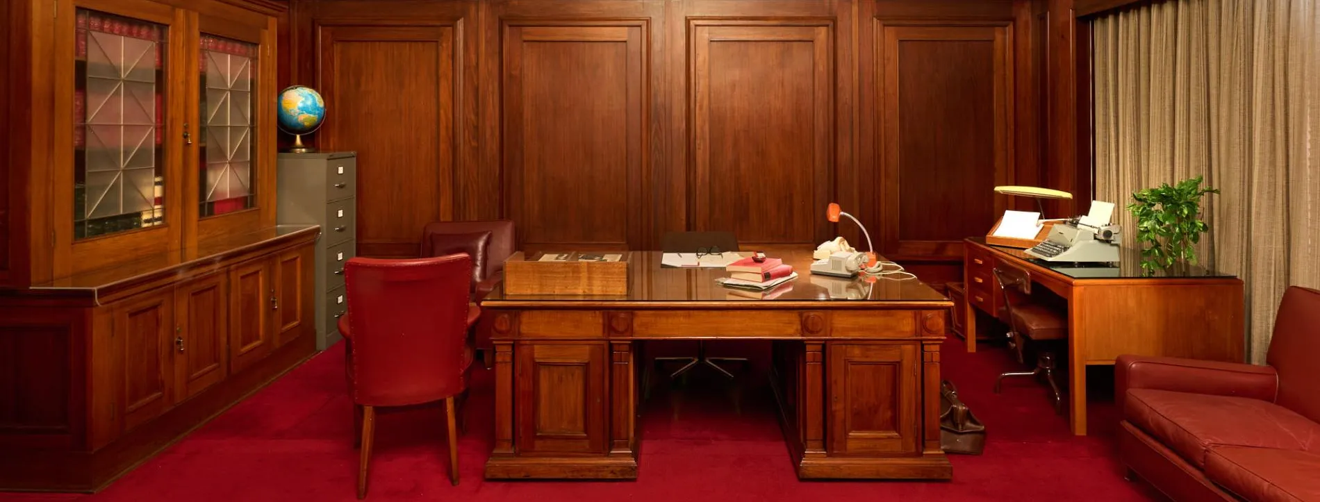 A wood-panelled room in Old Parliament House with two wooden desks, a wooden bookcase, red leather chairs and a red leather couch. The carpet is red.