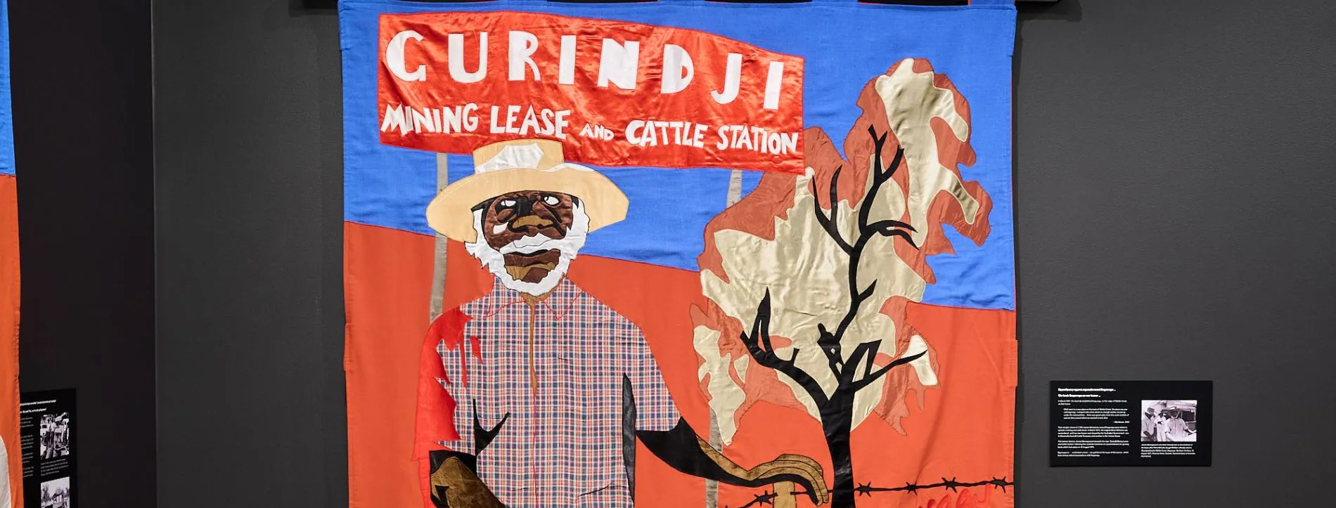 A Gurindji freedom banner depicting a First Nations person. The banner hangs in the exhibition on a black wall in Old Parliament House.