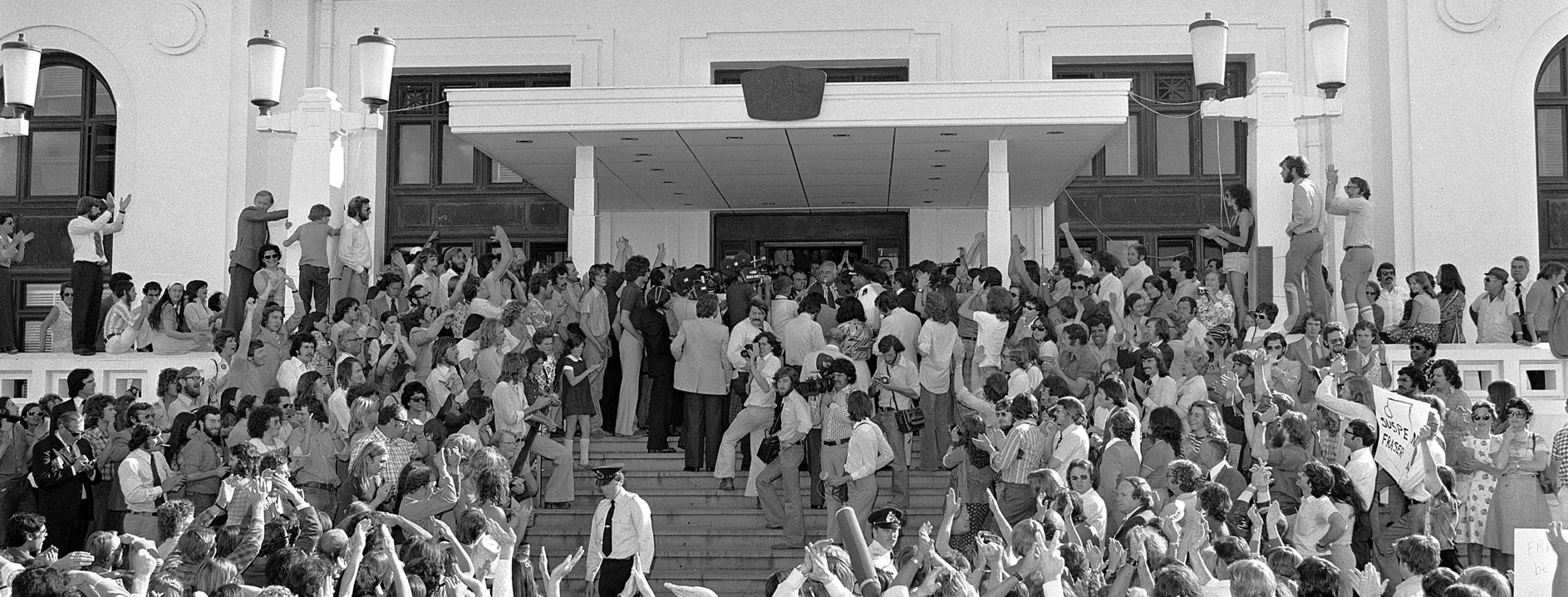 A large crowd of people and press stand below and on the steps of Parliament House after the dismissal of the Whitlam government.
