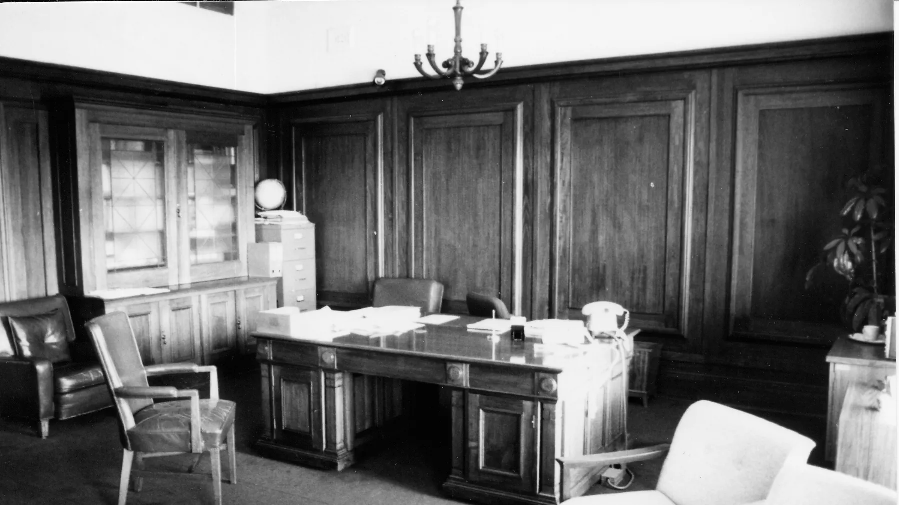 This black and white photograph shows the layout of the Clerk of the Senate’s office in 1985 when it was occupied by Alan Cumming Thom. The timber-panelled room contains a large desk (originally designed by the building’s architect, John Smith Murdoch), glass-fronted built-in bookshelves and a sideboard with an indoor plant. There are a range of visitor chairs and an easy chair in the foreground.