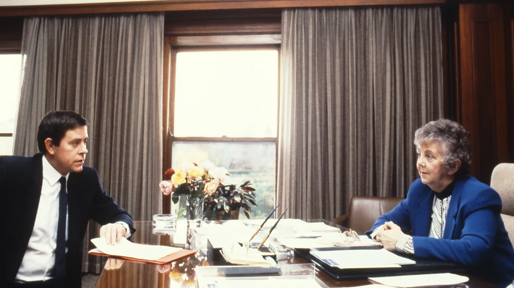 This professional colour photograph shows Principal Private Secretary John Porter and Speaker Joan Child sitting across from each other at the Speaker’s desk. John is wearing a dark suit, white shirt and dark tie and Joan is in a white blouse and vibrant blue jacket. They are discussing the daily program for the House of Representatives Chamber. There is a vase of roses, stationery and paperwork on the desk. In the background the curtains have been drawn to reveal a large east-facing window.