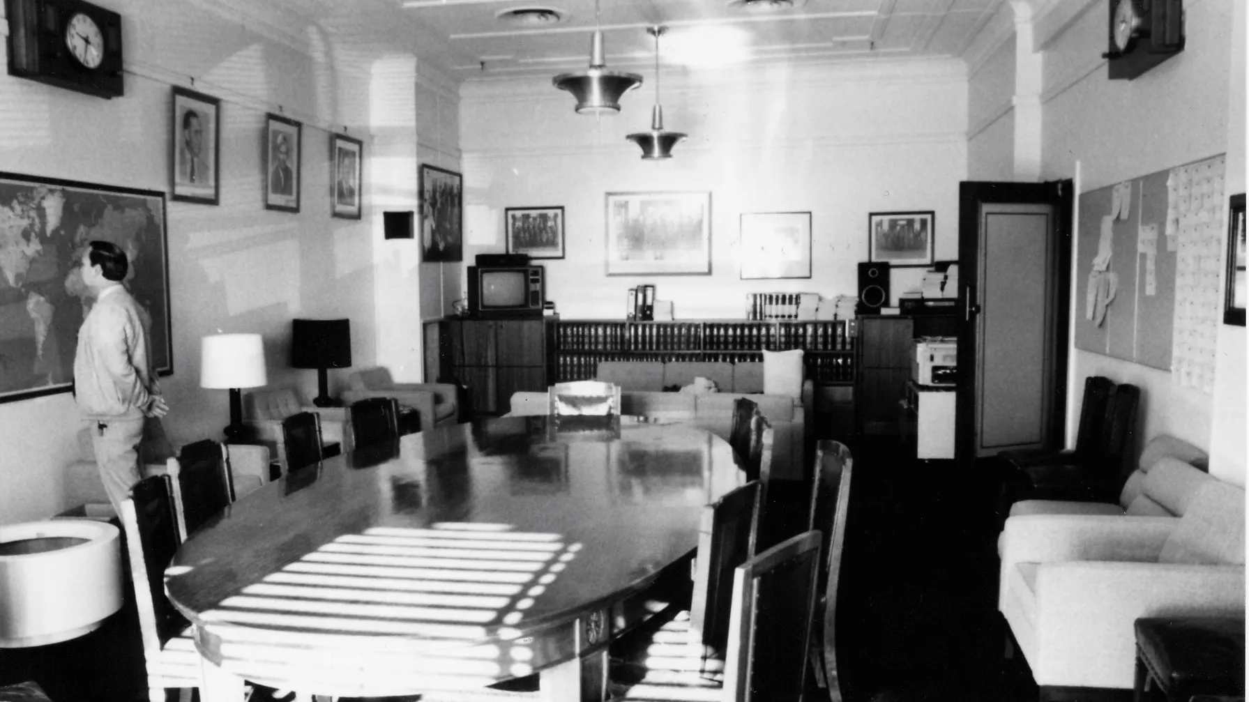 A large oval meeting table and chairs dominate the centre of the room. Easy chairs with side tables and lamps are arrayed along the sides and two sofas are arranged in a seating area towards the back of the room. A sideboard with books, a television and sound system stretches along the back wall. There is a framed map on one wall and a notice board on the opposite. Portraits of leaders and formal and informal photographs of the party crowd the walls