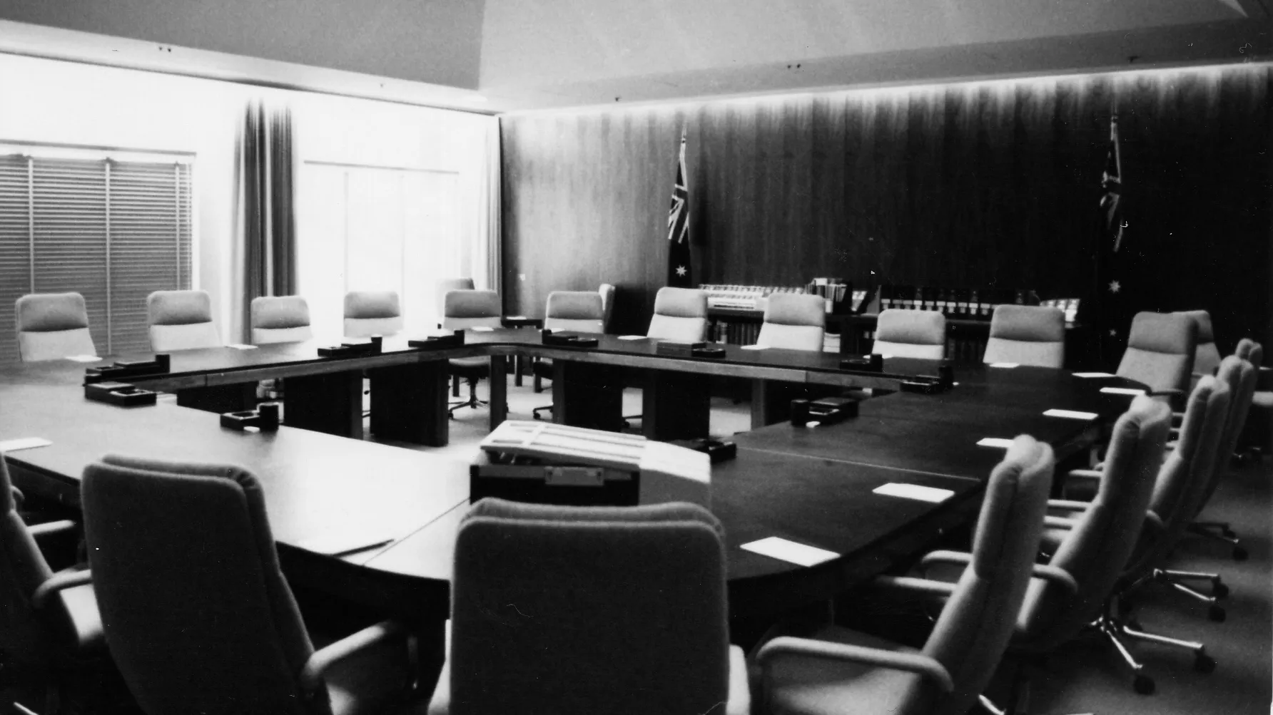 The Cabinet Room at Old Parliament House with a square table surrounded by chairs. Two Australian flags stand along a wood panelled wall.
