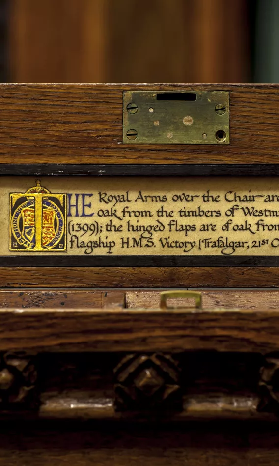 Cursive script on a label on the side of the Speaker's Chair says 'The Royal Arms over the Chair are carved in oak from the timbers of Westminster Hall (1399); the hinged flaps are of oak from Nelson's flagships HMS Victory (Trafalgar, 21st October, 1805)'.