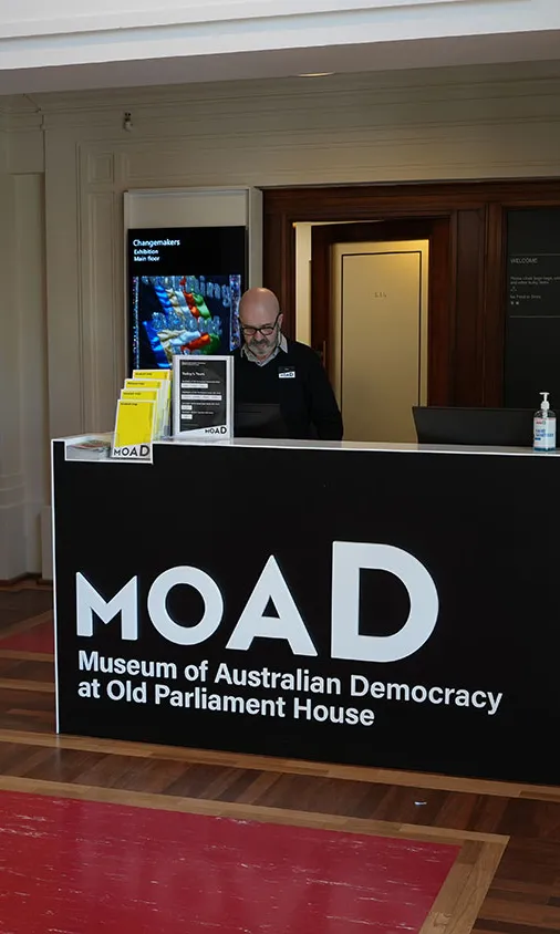 A museum staff member wearing a black sweater and name badge stands behind the main entrance reception desk at MoAD, the Museum of Australian Democracy at Old Parliament House.